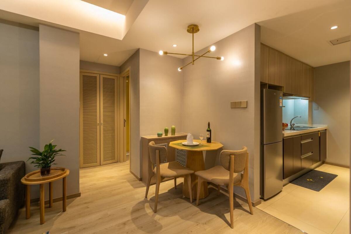 A modern kitchen with a built-in oven, a refrigerator, and a sink. There is a dining area with a table and chairs, and a small plant on a side table.