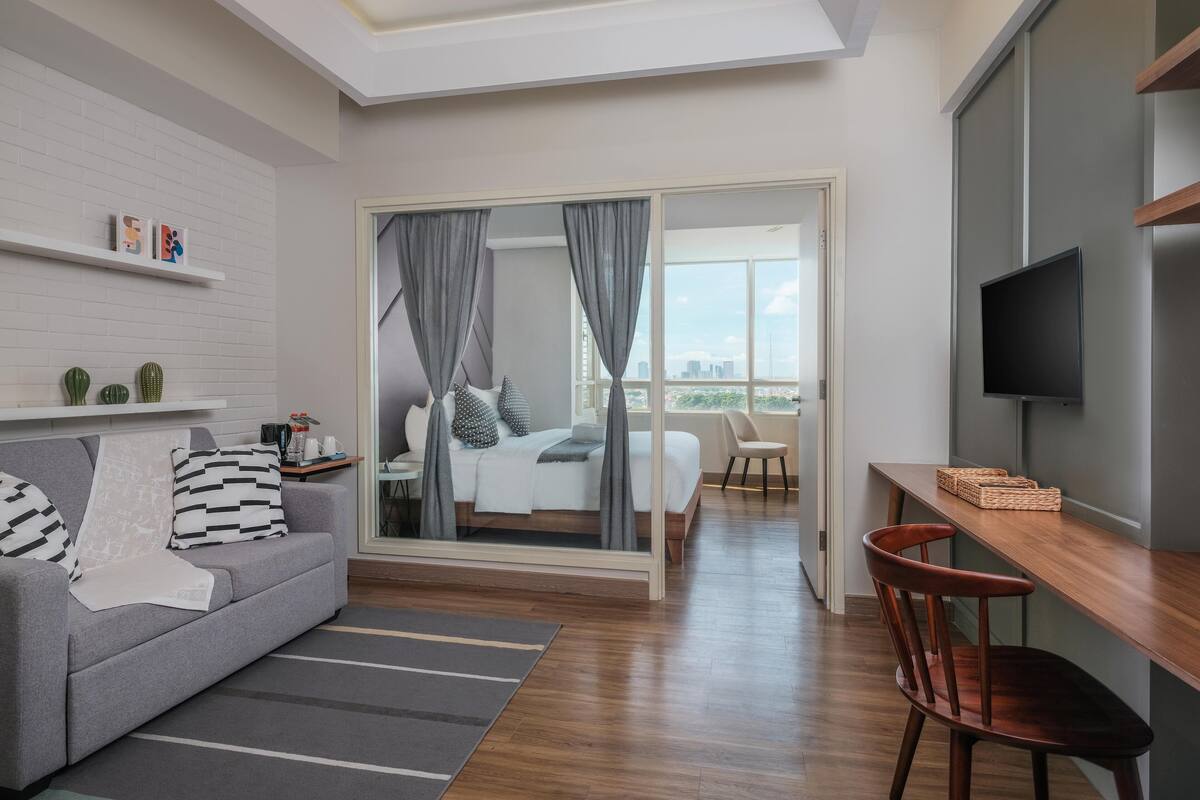 A modern hotel room with a sofa, a dining table, a TV, and a bed with a city view.