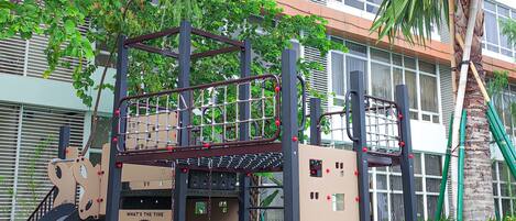 A playground with climbing structures and a slide, surrounded by a building with large windows and a palm tree.