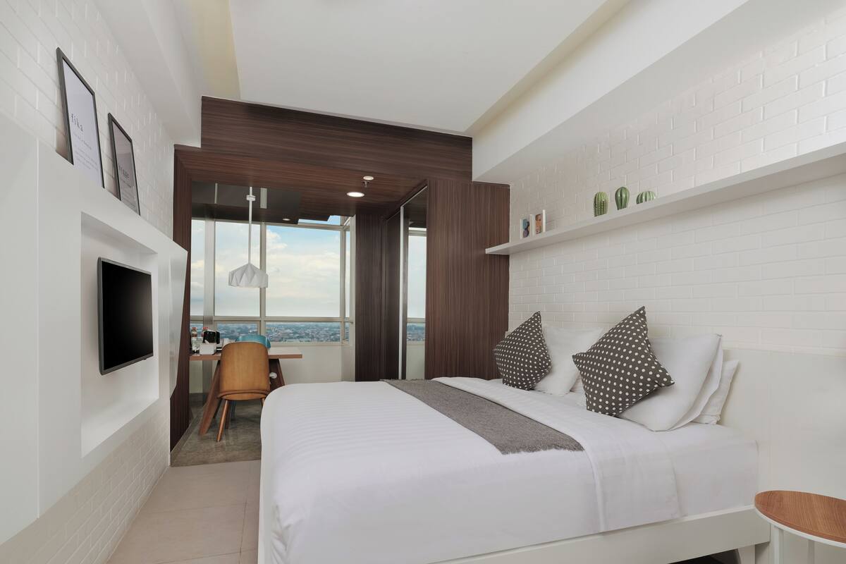 A modern hotel room with a large bed, a TV mounted on the wall, a desk with a chair, and a view of the city through large windows.
