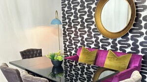 A dining area with a glass table, four upholstered chairs, a potted plant, a blue floor lamp, and a patterned wall with a gold mirror.