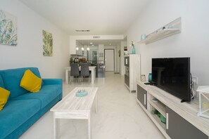 A modern living room with a blue sofa, a coffee table, a TV on a wooden stand, and a kitchen in the background.