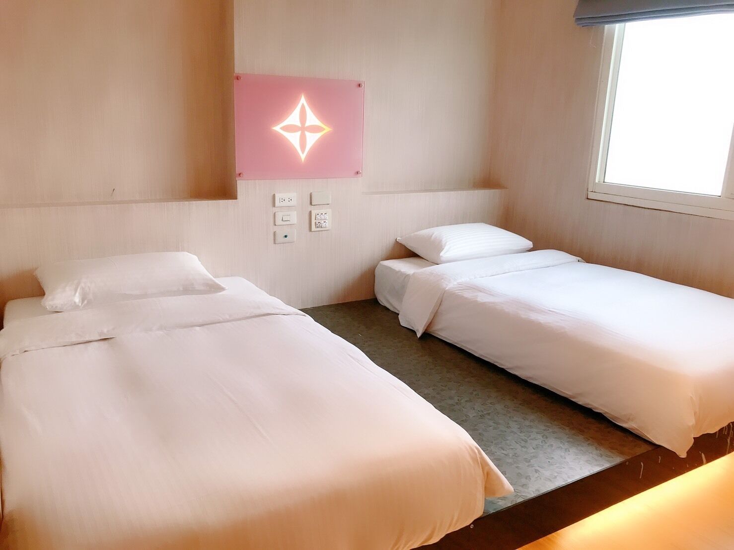 A hotel room with two single beds, a wall-mounted light fixture, and a window.