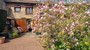 Un bâtiment en pierre avec une porte et des fenêtres en bois, entouré d’une cour en gravier et d’arbres en fleurs.