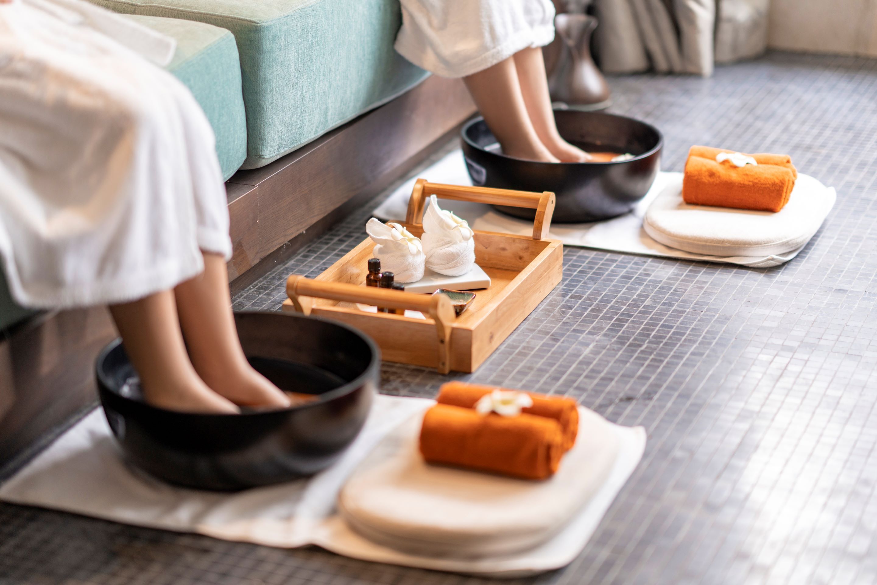 A spa setting with a wooden tray holding towels and a bowl, a person sitting in a black bowl, and a foot bath with a foot in a bowl.