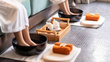 A spa setting with a wooden tray holding towels and a bowl, a person sitting in a black bowl, and a foot bath with a foot in a bowl.