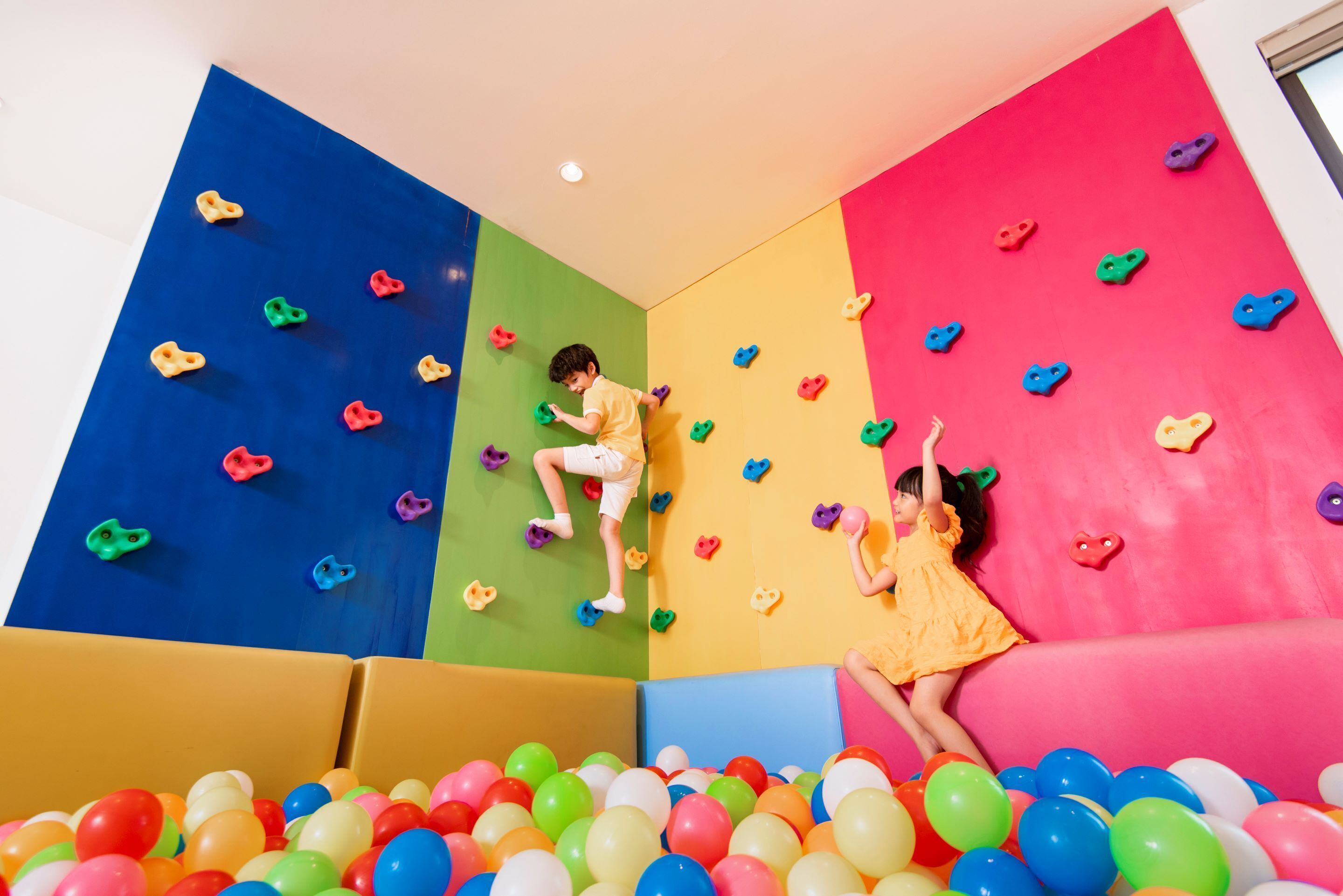 Un enfant escalade un mur d’escalade intérieur coloré, avec une piscine à balles au pied du mur.