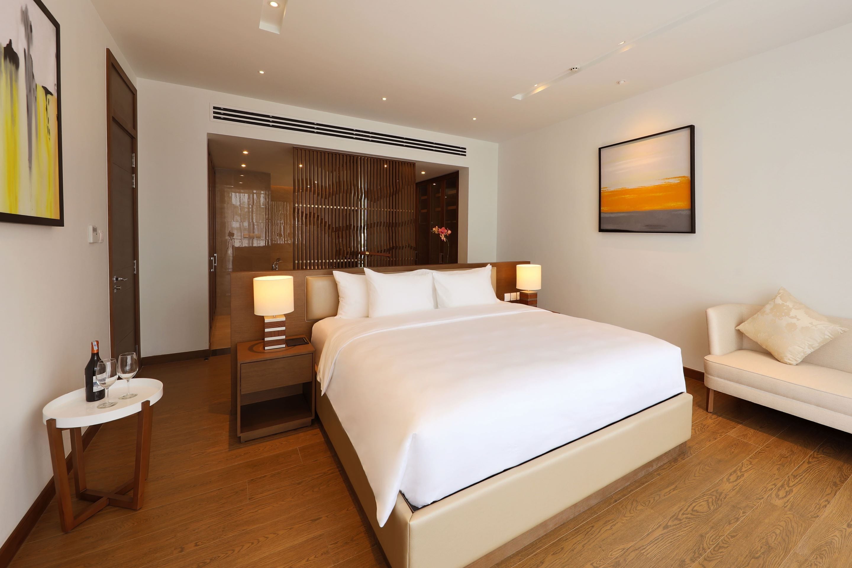 Une chambre d’hôtel moderne avec un grand lit, des tables de chevet, une petite table ronde avec une bouteille et des verres, un canapé et un tableau au mur.