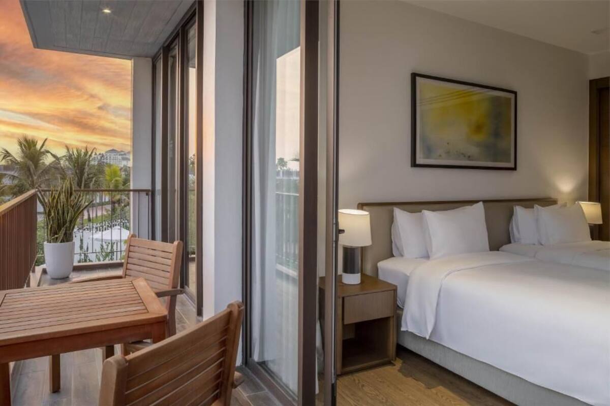 Habitación de hotel con balcón, cama, mesita de noche y un cuadro en la pared.