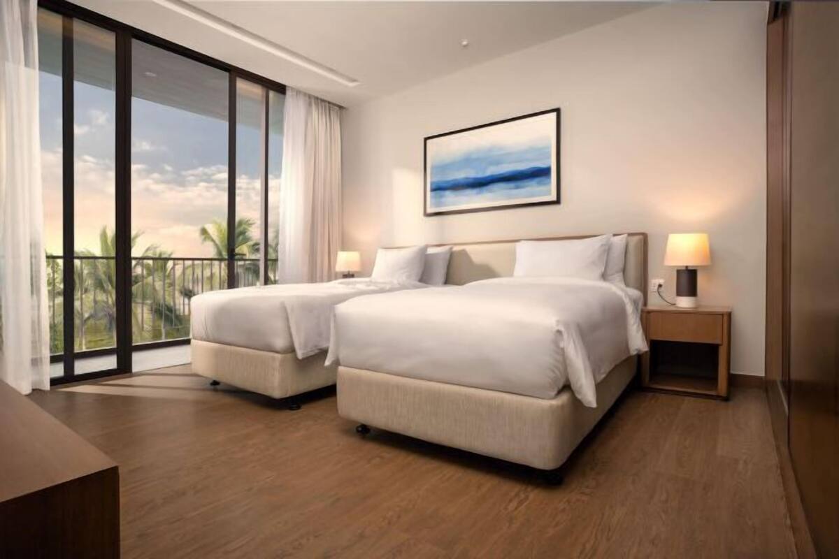 Habitación de hotel con dos camas, un ventanal con vistas, un cuadro enmarcado en la pared y una mesita de noche con una lámpara.