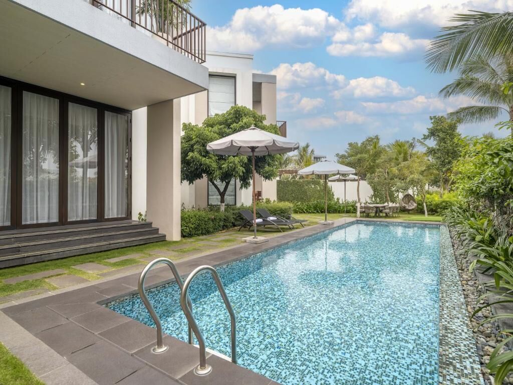 Un espace piscine moderne avec une piscine à l’eau d’un bleu limpide, des chaises longues, des parasols et un bâtiment doté de grandes fenêtres et d’un balcon.