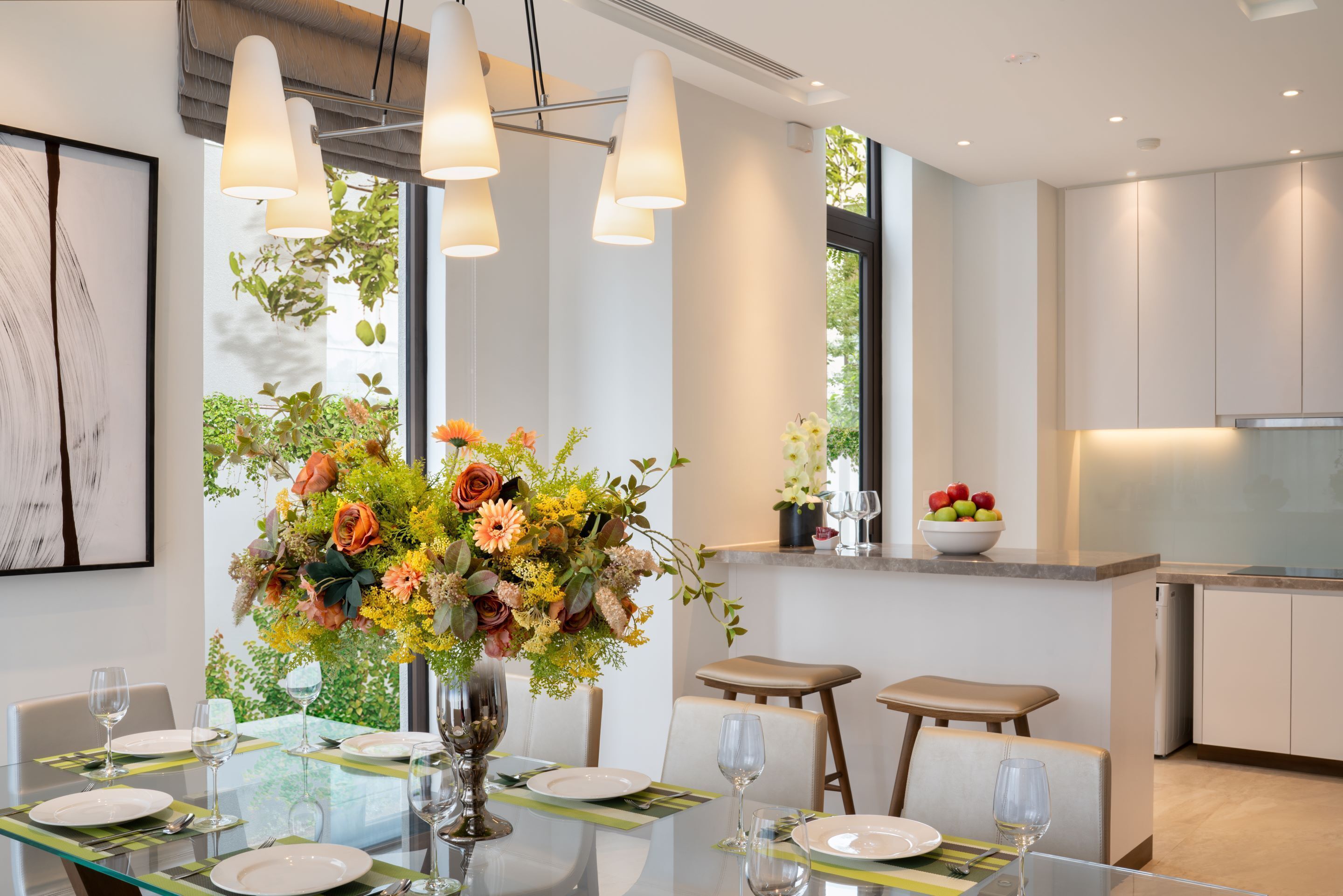 Un comedor moderno con una mesa de vidrio, sillas y un centro de mesa con flores. Se ve un área de cocina con armarios y una encimera al fondo.