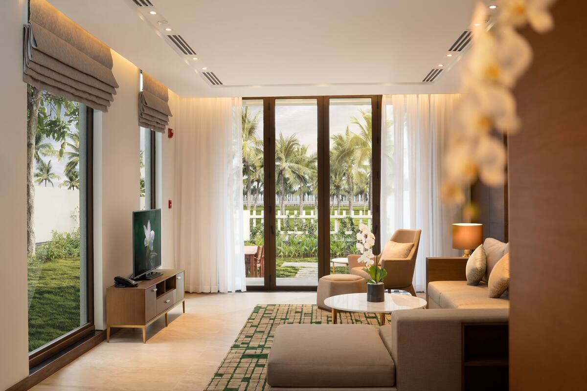 Una sala de estar moderna con sofá, mesa de centro y televisor, ventanas amplias con cortinas y vista a palmeras.