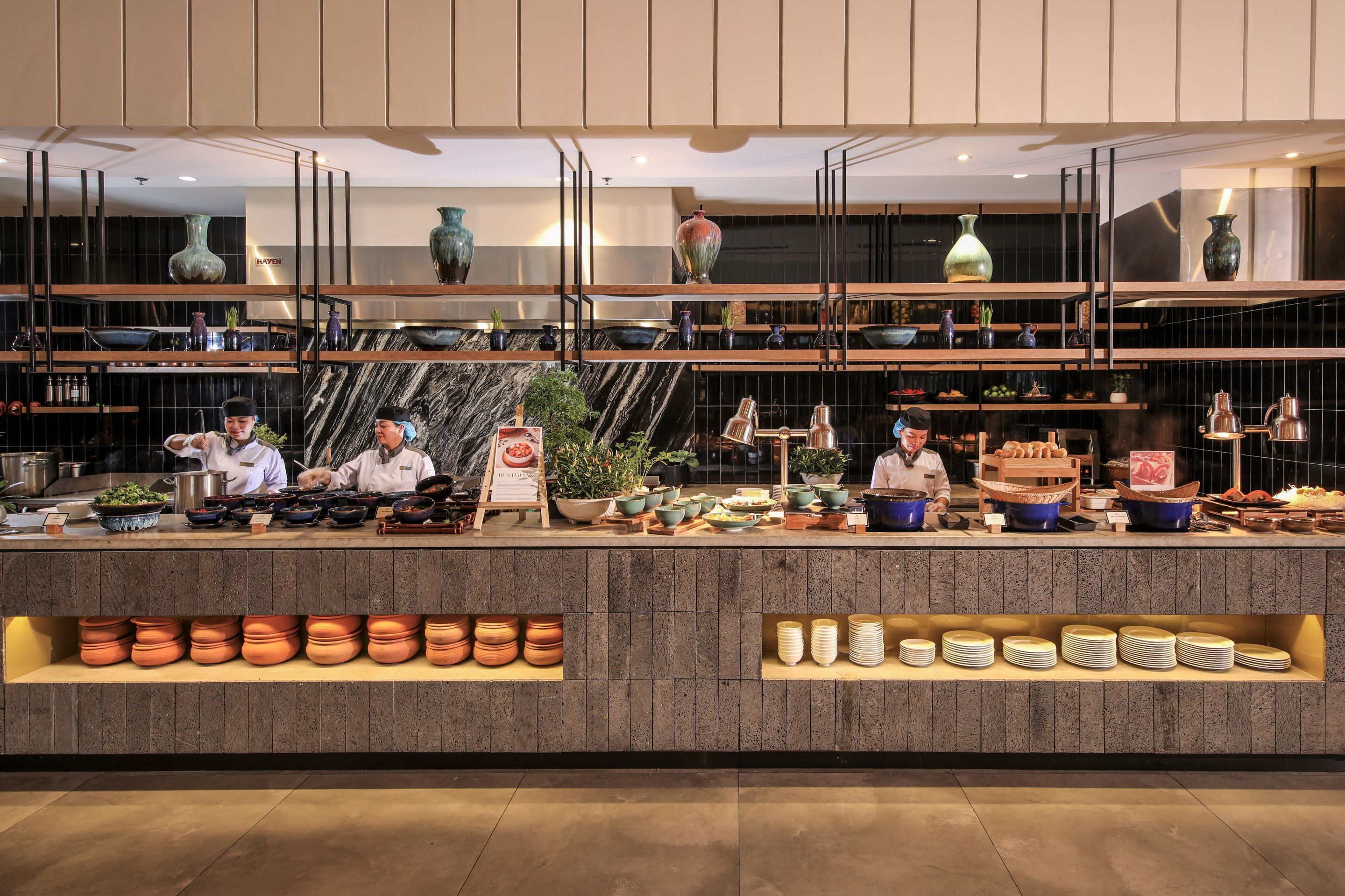 A modern kitchen with chefs preparing food, shelves with kitchenware, and decorative vases.