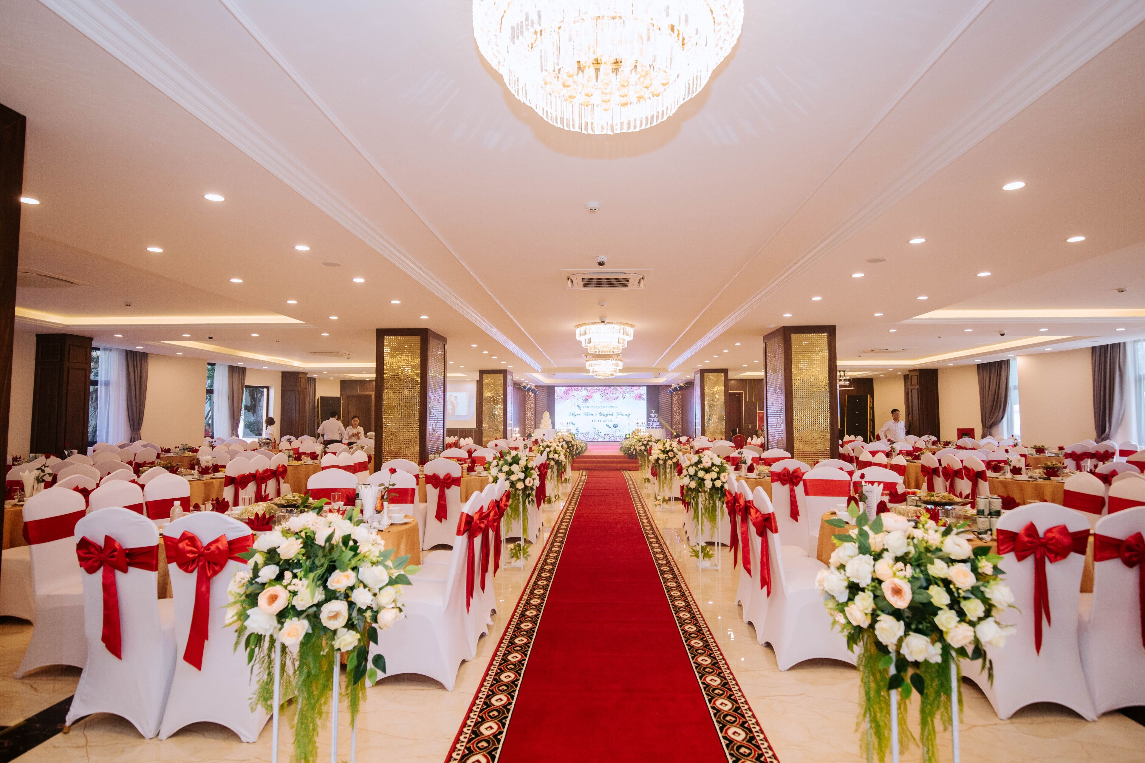 Un salón de banquetes con filas de sillas blancas adornadas con cintas rojas, un pasillo con alfombra roja y una lámpara de araña.