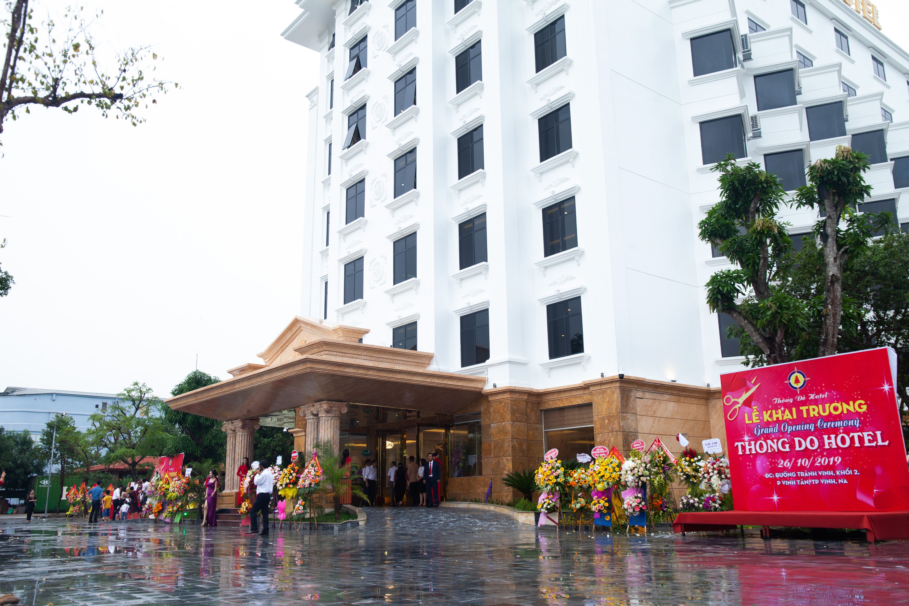 A hotel entrance with a grand opening ceremony, featuring a large red sign, a fountain, and people gathered outside.