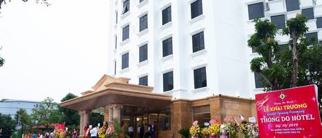 Entrada de hotel con una ceremonia de inauguración, que incluye un gran letrero rojo, una fuente y personas reunidas afuera.