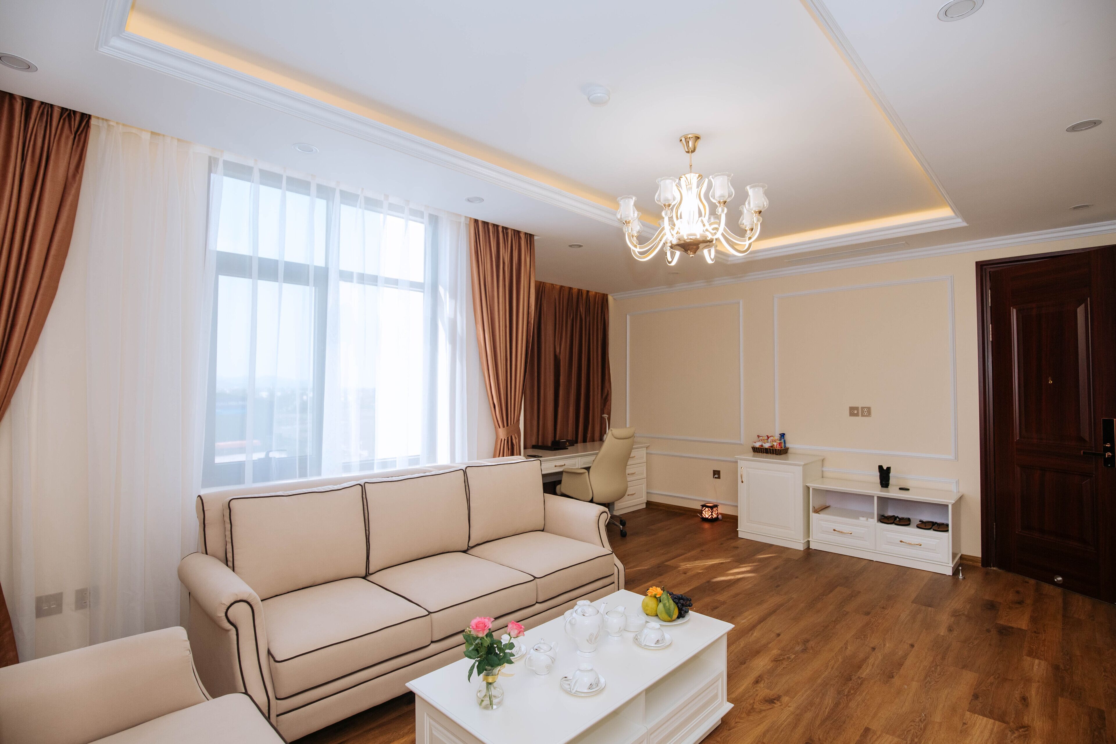 A modern living room with a beige sofa, a white coffee table with decorative items, a desk, and a chandelier.