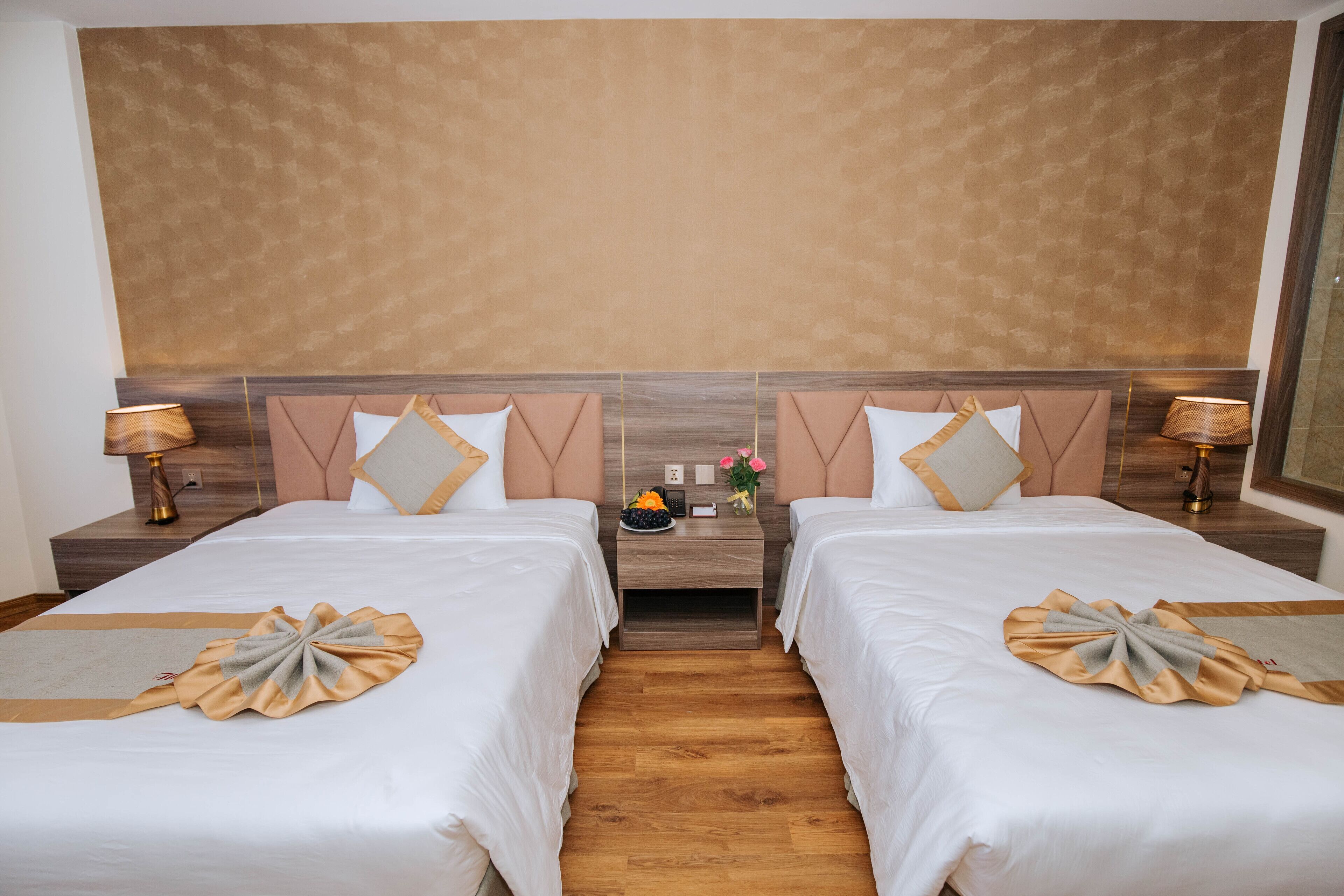 A hotel room with two beds, wooden headboards, bedside tables with lamps, and a small table with a vase of flowers.