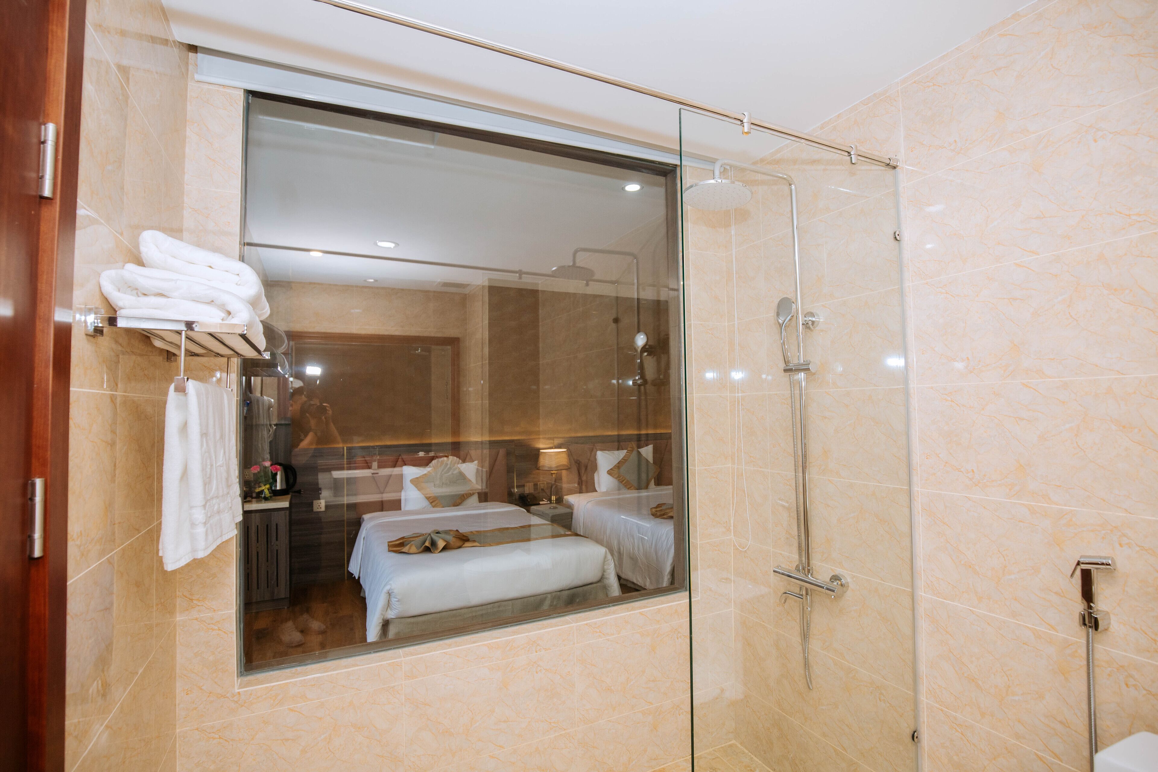 A modern bathroom with a glass shower enclosure, a visible bed with a bench, and a towel rack with folded towels.
