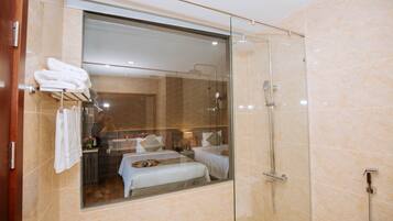 A modern bathroom with a glass shower enclosure, a visible bed with a bench, and a towel rack with folded towels.