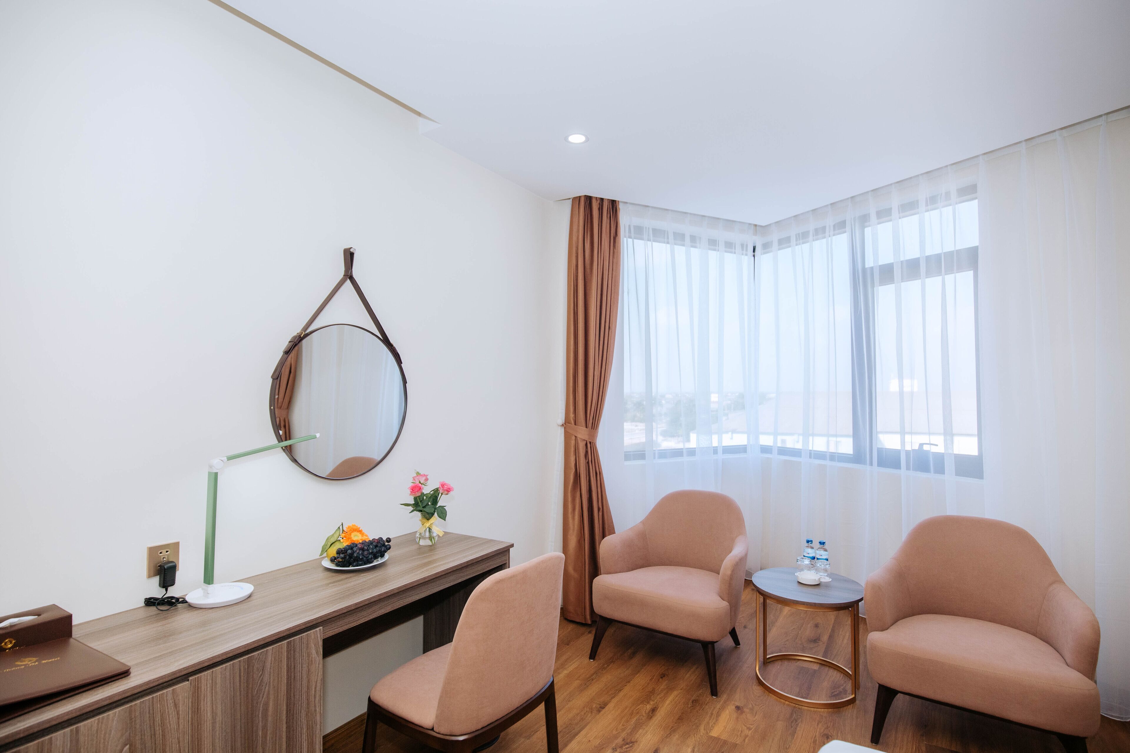 A hotel room with a wooden desk, two armchairs, a small round table, a mirror, and a window with sheer curtains.