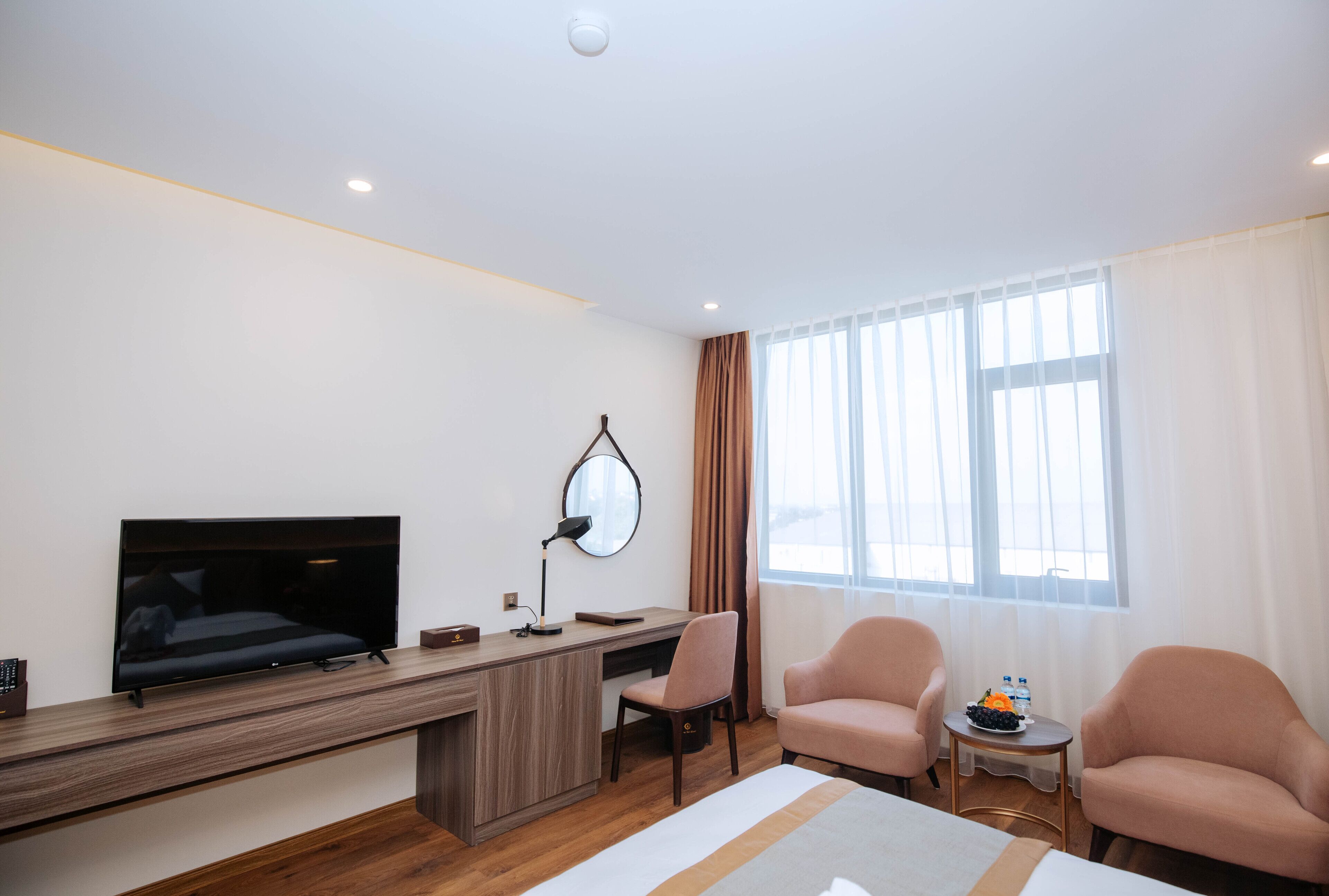 Una habitación de hotel moderna con un escritorio de madera, un televisor de pantalla plana, un sillón cómodo, una pequeña mesa redonda con frutero y un ventanal grande con cortinas translúcidas.