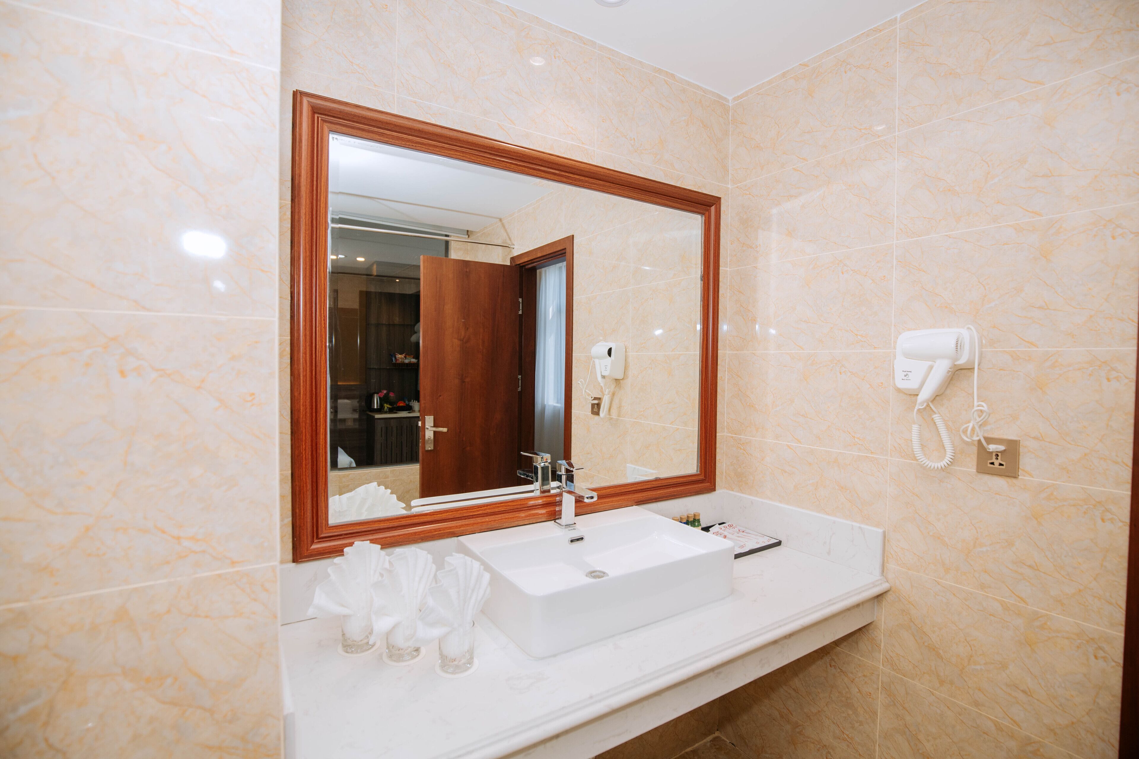 A bathroom with a marble wall, a wooden-framed mirror, a white sink, and a hairdryer on the wall.