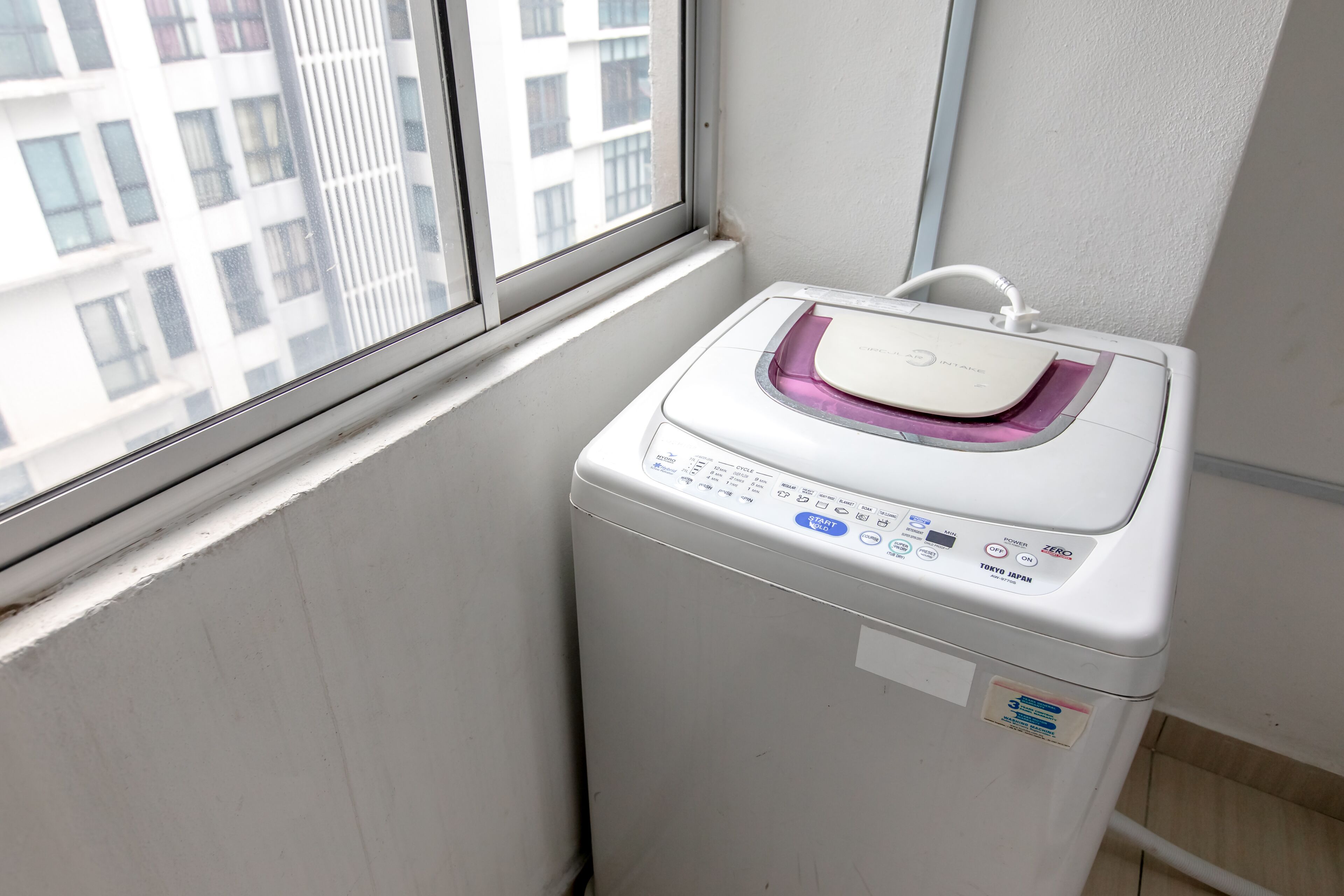 Un lave-linge compact, de couleur blanche, doté d’un panneau de commande, placé près d’une fenêtre donnant sur un bâtiment.