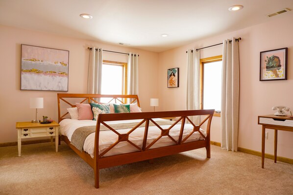 A bedroom with a wooden bed, two bedside tables, a nightstand, a desk, and two windows with curtains.