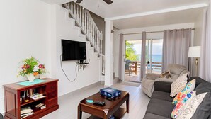 A living room with a staircase, a flat-screen TV, a coffee table, a sofa, and a view of the ocean. - Nautilus One – A Beachfront Condo on Seven Mile Beach - by Cayman Villas (West Bay)