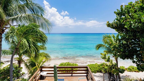 A wooden deck with lounge chairs overlooking a beach with clear turquoise water and palm trees. - Nautilus One – A Beachfront Condo on Seven Mile Beach - by Cayman Villas (West Bay)