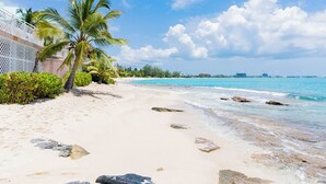 A beach with white sand, palm trees, and clear blue water. - Nautilus One – A Beachfront Condo on Seven Mile Beach - by Cayman Villas (West Bay)