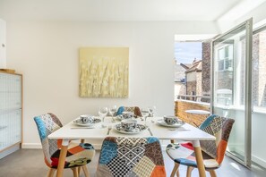 Zona de menjador amb taula preparada per a quatre, cadires amb tapisseria estampada, un quadre abstracte groc a la paret i vista d'un edifici de maó a través d'una porta de vidre.