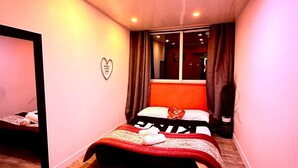 Un dormitorio con cama, una silla, una decoración con forma de corazón en la pared y una ventana con cortinas.