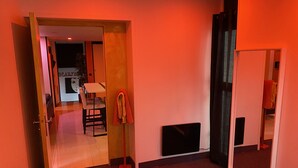 Un pasillo con alfombra roja, una puerta que da a otra habitación, una toalla roja en un perchero y un objeto negro en la pared.