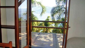 A room with a bed, a small table, and a balcony with a view of palm trees and mountains.