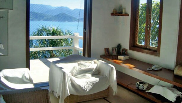 A room with a bed, a small table, and a window with a view of the sea and mountains.