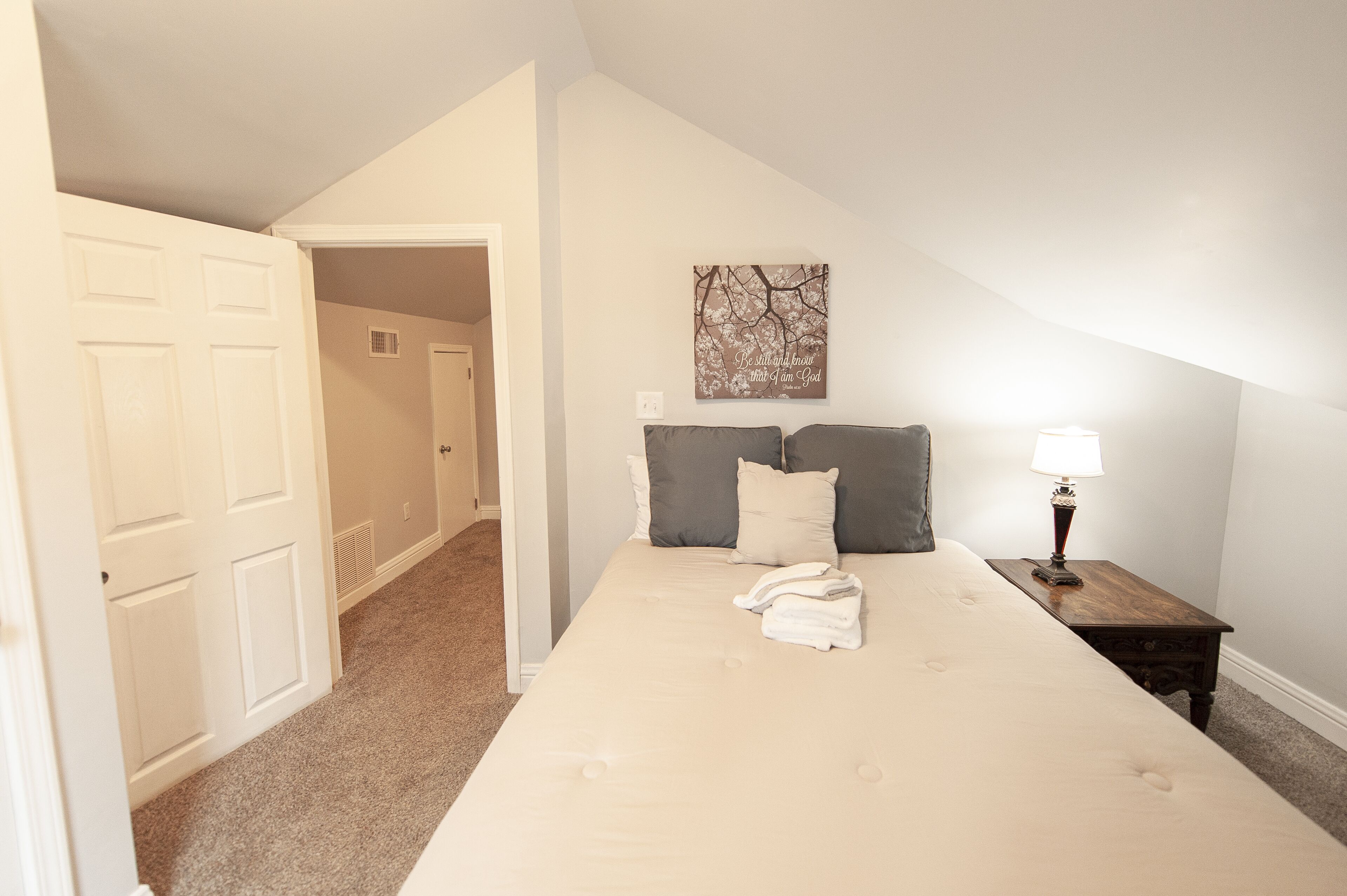 Une chambre avec un grand lit, une table de chevet avec une lampe, un tableau décoratif au mur et une porte donnant sur une autre pièce.