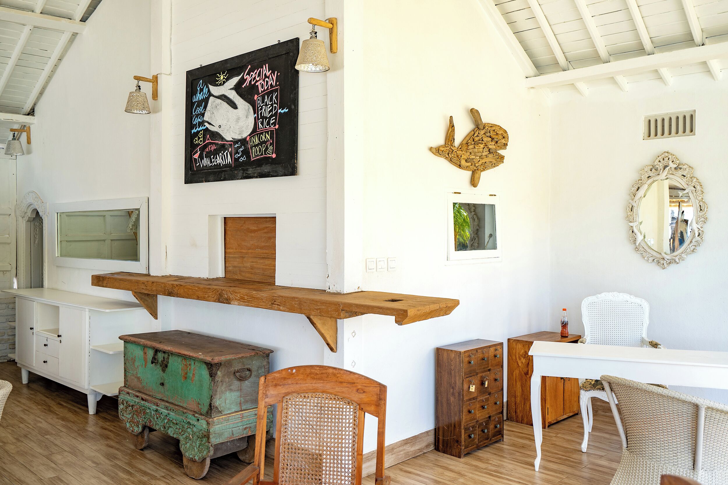 Ein Zimmer mit einem hölzernen Regal, einem weißen Schreibtisch, einem Korbstuhl und einem Spiegel an der Wand.