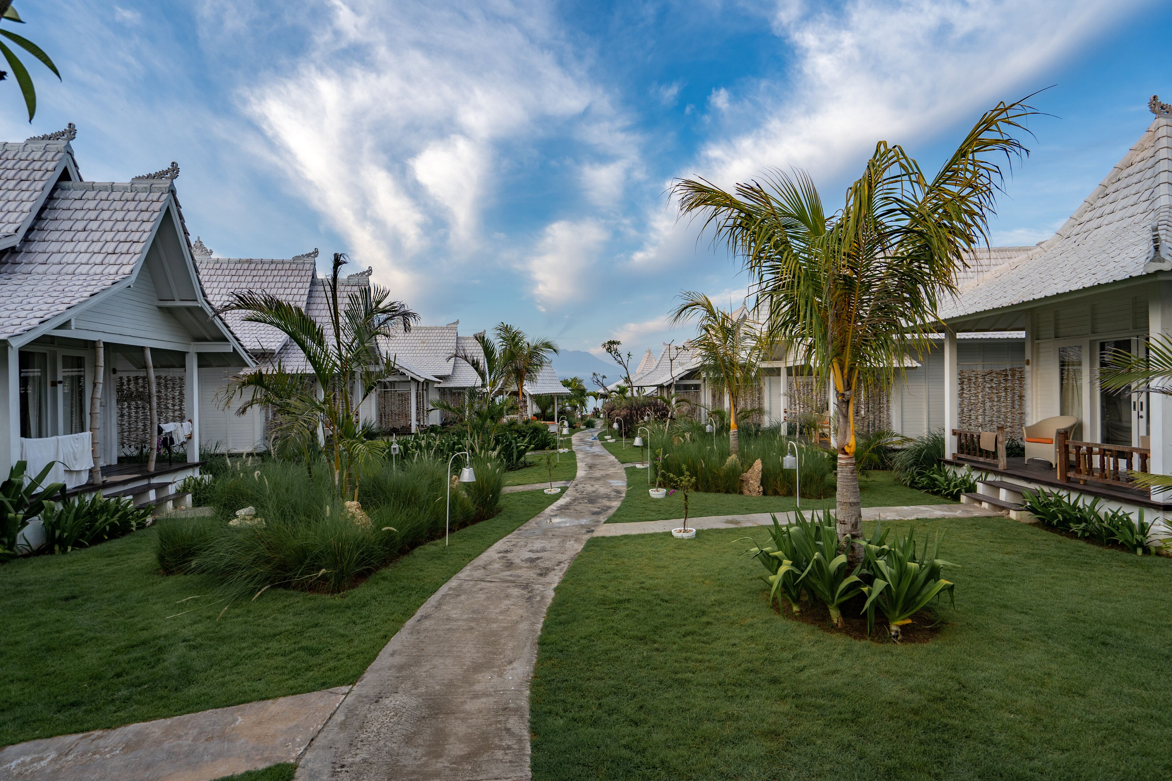 A pathway leading through a well-maintained garden with palm trees and various plants, surrounded by traditional-style buildings with tiled roofs.
