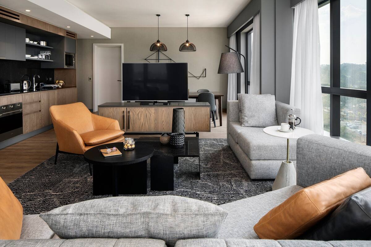 Uma sala de estar moderna com sofá, poltrona e mesa de centro. Há uma TV fixada na parede e uma área de cozinha com armários e eletrodomésticos.