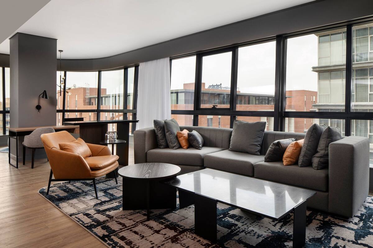 Uma sala de estar moderna com um sofá cinzento, uma poltrona laranja e uma mesa de centro. Janelas amplas oferecem vista para a paisagem urbana.