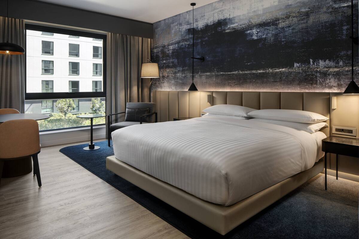 Quarto de hotel moderno com uma cama grande, uma escrivaninha e vista para um edifício pela janela.