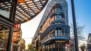 An outdoor seating area with wooden stools and a curved railing, overlooking a modern building with a curved facade and a sign reading "Mr. George." - Johannesburg Marriott Hotel Melrose Arch (Johannesburg)