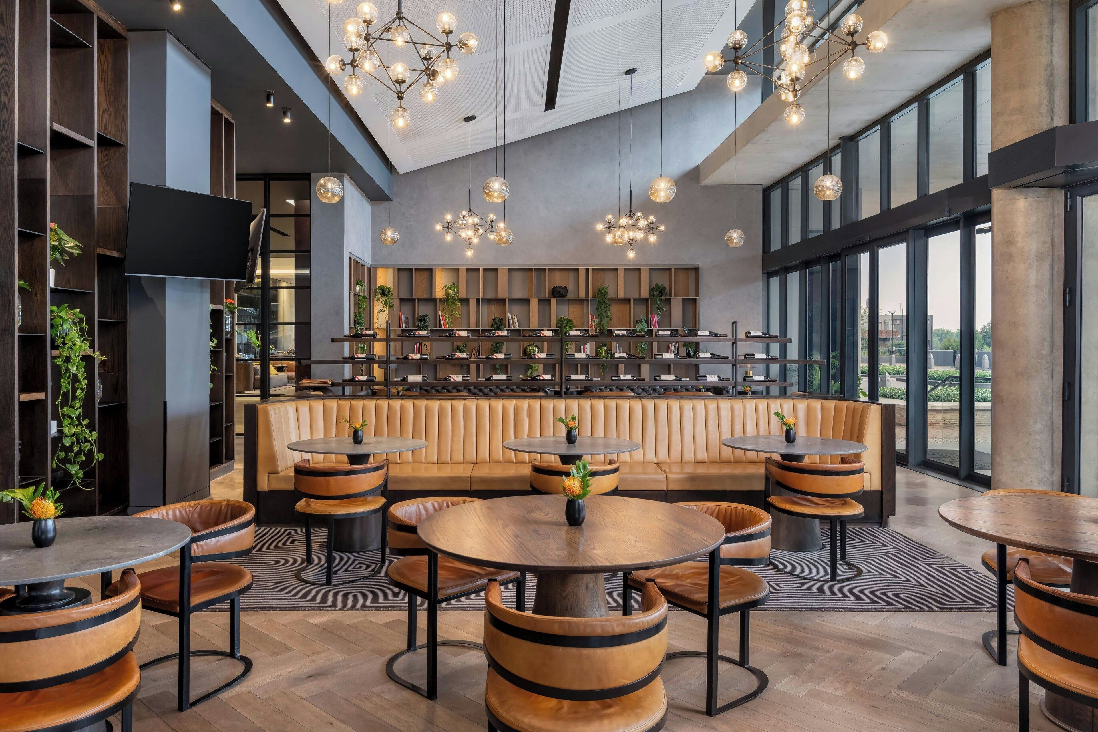 A modern interior with round tables and leather seating, a large TV, and a bar area with shelves and plants.