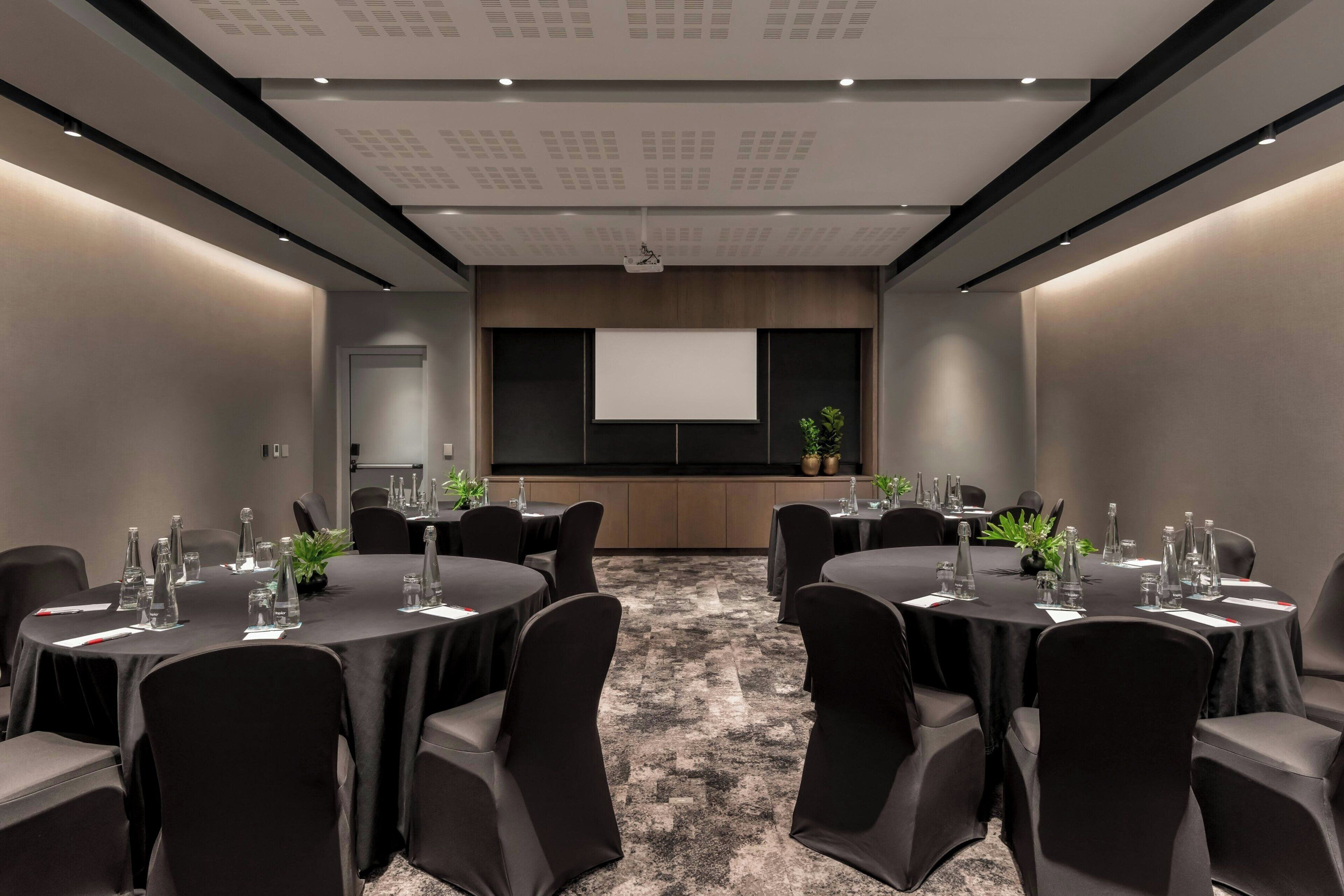 A conference room with round tables, black chairs, and a projector screen.