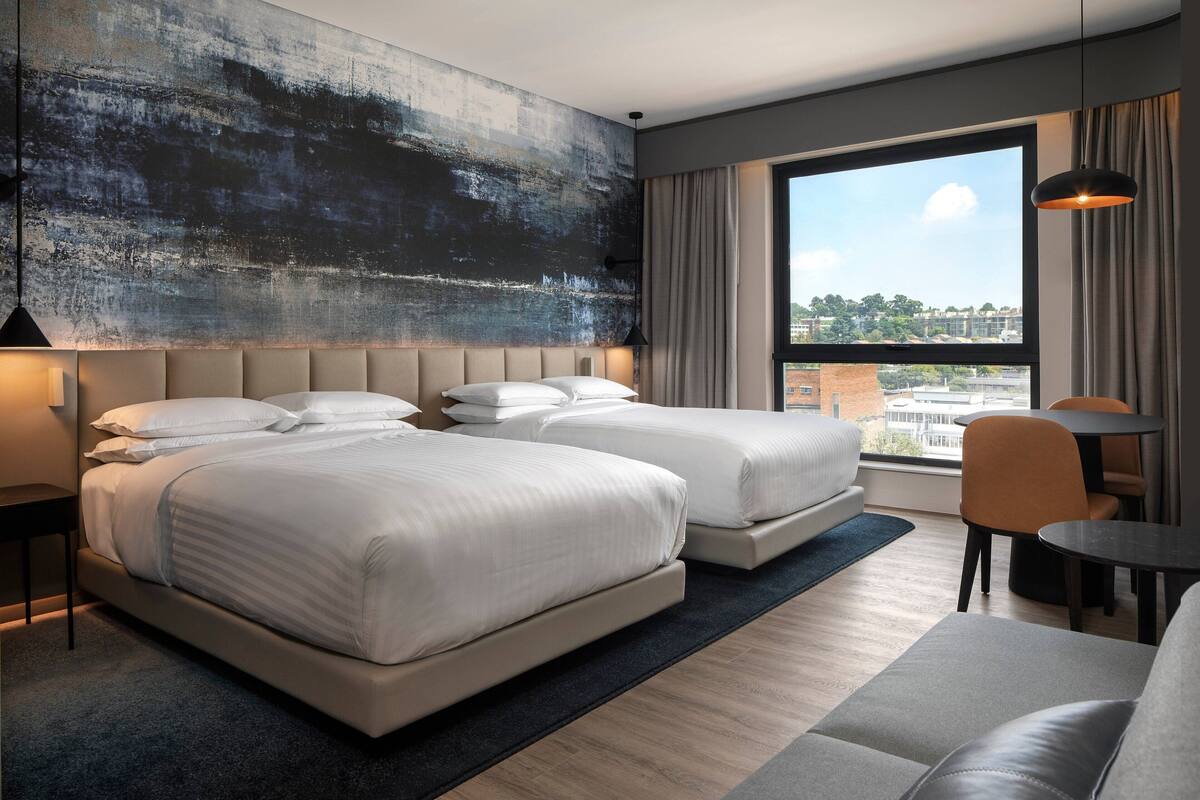 Quarto de hotel moderno com duas camas, uma janela ampla com vista para a cidade, um sofá e uma mesa pequena.