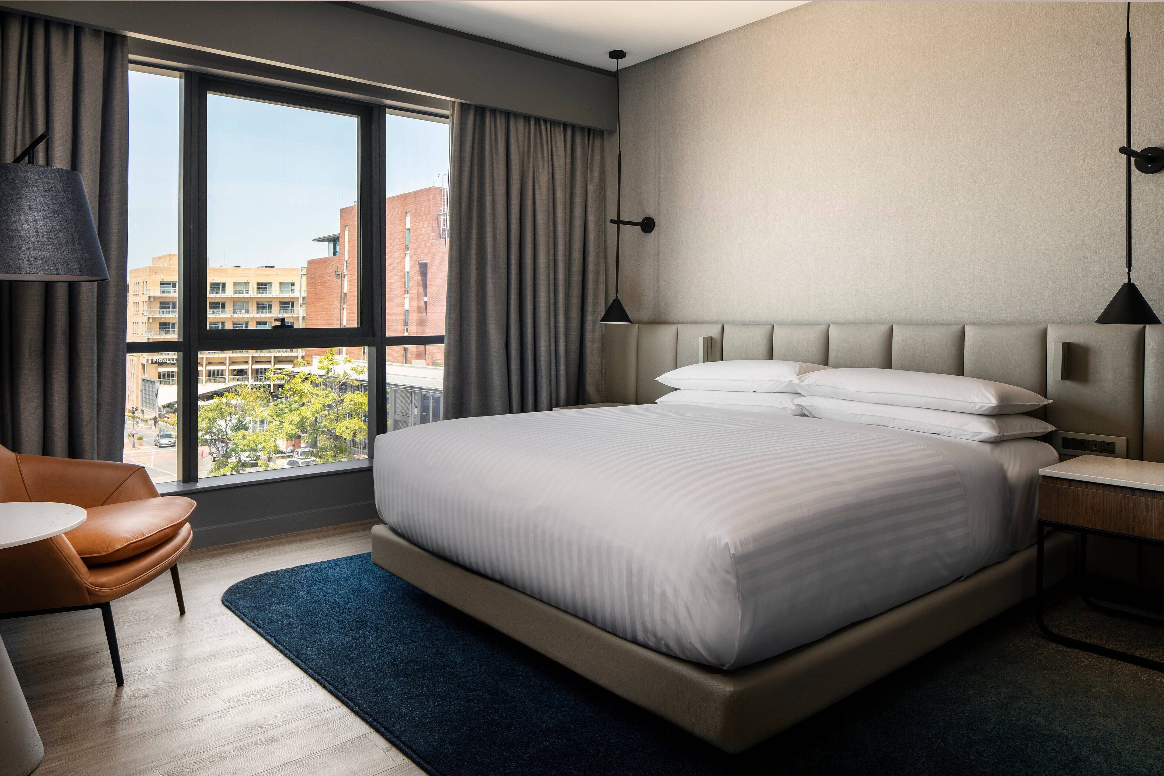 Ein modernes Hotelzimmer mit einem großen Bett, einem Schreibtisch, einem Sessel und Blick auf die Stadt durch große Fenster.