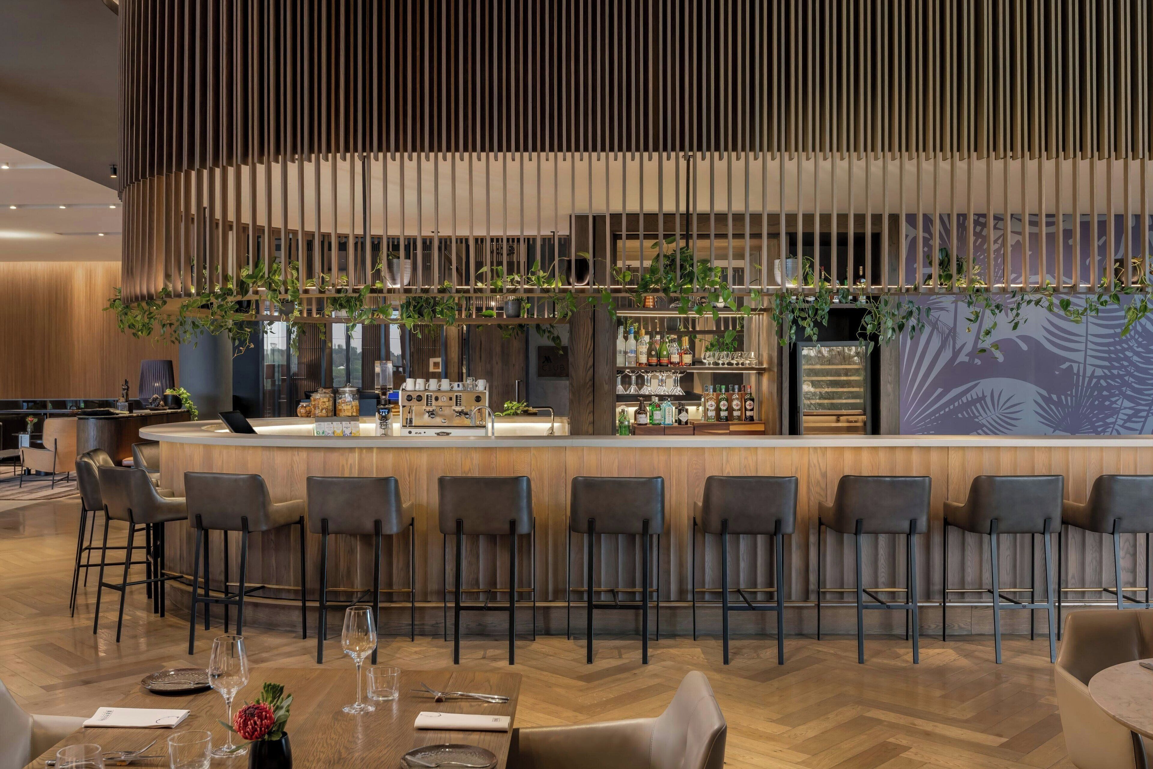 Zona de bar moderna con mostrador de madera, taburetes y una variedad de botellas y vasos.